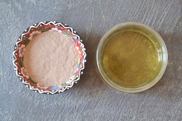 Top view of dry clay (rhassoul, ghassoul) in a colorful porcelain bowl with apple cider vinegar on a gray stone background.