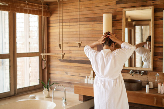 Woman In White Dressing Gown Standing In A Bathroom Tying Her Hair