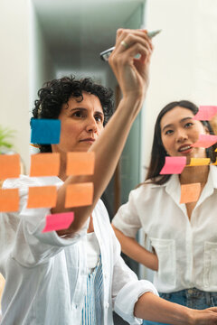 Multiracial businesswomen putting post-it notes on glass board