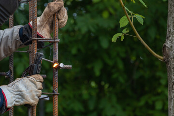 A worker welds a reinforcement - future column - construction concept. Green trees background