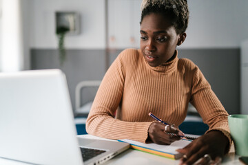 Black woman writing in notepad while working on laptop