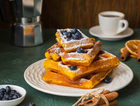 Stacked Pumpkin Waffles, sprinkled with powdered sugar, with fresh blueberries on a white plate on a green concrete background. Waffle recipes. Pumpkin dishes. Thanksgiving day