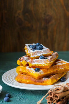 Stacked Pumpkin Waffles, sprinkled with powdered sugar, with fresh blueberries on a white plate on a green concrete background. Waffle recipes. Pumpkin dishes. Thanksgiving day