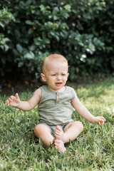 Not happy baby boy in a green outfit sitting on the grass at a park