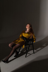 Obraz na płótnie Canvas fashion model girl shooting in a mustard-colored dress on a white background on a cyclorama sitting on a chair subdued light 