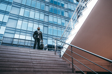 Obraz na płótnie Canvas An elegant African man entrepreneur is standing on the stairs of a yard of a business skyscraper; a black businessman in a fashionable outfit with a bag on the stairway of an office high-rise