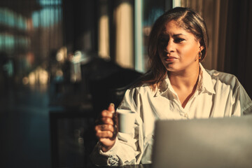 Portrait of a ravishing Hispanic female drinking coffee while using her laptop in a cafe; beautiful adult plus size Brazilian businesswoman having a coffee break; with a copy space place on the left - obrazy, fototapety, plakaty