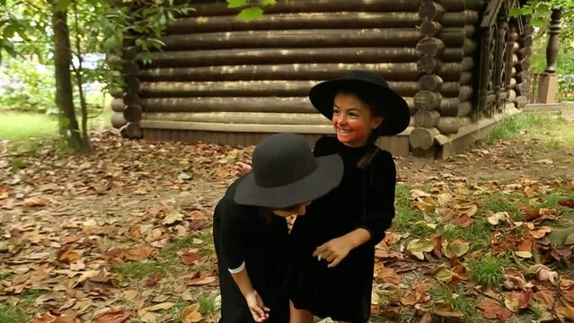 Two little girls, in black witch costumes, and hats, paint each other with scary makeup and laugh, on Halloween
