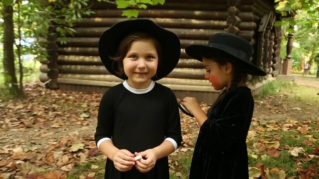 Two little girls, in black witch costumes, and hats, paint each other with scary makeup and laugh, on Halloween
