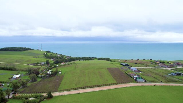 Vista a&eacute;rea desde dron por un campo verde que llega hasta la playa en donde se ven casas, caba&ntilde;as, el mar y sus olas
