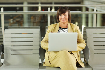 Mature businesswoman is sitting at the airport in terminal hall with a laptop and looking in camera. Middle ages female is waiting for departure at the gate, working online. Freelance. copy space.