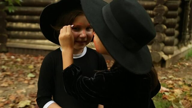 Two little girls, in black witch costumes, and hats, paint each other with scary makeup and laugh, on Halloween
