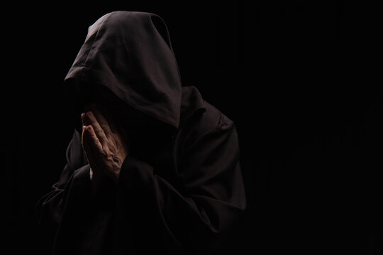 mysterious priest in hooded robe praying isolated on black.