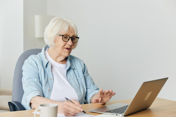 portrait of an elderly woman in a light shirt working at a desk through a laptop at home, carefully looking at the monitor. The concept of working from home