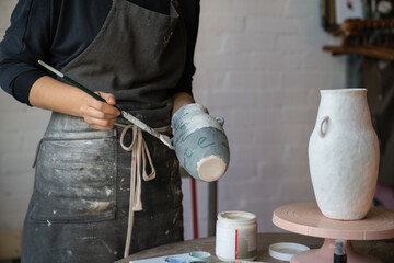 Woman in apron works with pottery standing near round wooden table with equipment. Professional...