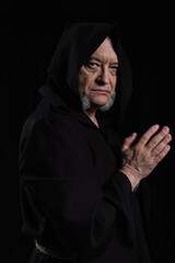 strict monk in hooded cassock looking at camera while standing with praying hands isolated on black.