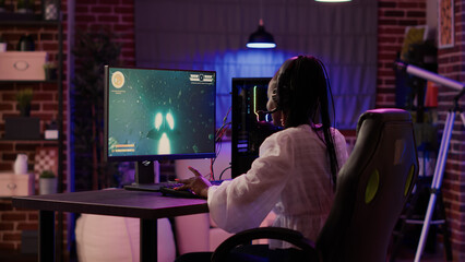 Woman playing multiplayer space shooter simulation enjoying free time using pc gaming setup in home living room. African american gamer girl talking in headset while streaming online action game.