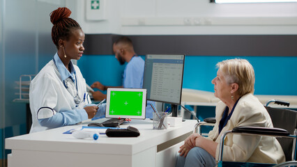 Obraz premium Physician holding tablet with horizontal greenscreen at appointment with woman in wheelchair. Using chroma key display with isolated copyspace and blank mockup background. Tripod shot.