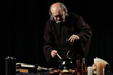 medieval alchemist preparing magic potion while adding ingredient into pot isolated on black.