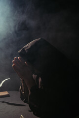 monk with obscured face praying with rosary at night on black with smoke.