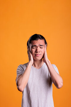 Young asian man covering ears with hands, hear no evil, three wise monkeys moral concept. Scared teenager with closed eyes showing no noise gesture, front view studio medium shot