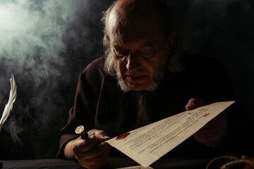 senior priest looking at wax seal on manuscript on dark background with smoke.