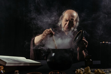 senior alchemist frowning near steaming pot while cooking at night on black background.