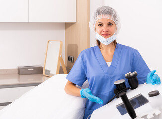 Portrait of positive female doctor cosmetologist wearing gloves and blue overall showing equipment...