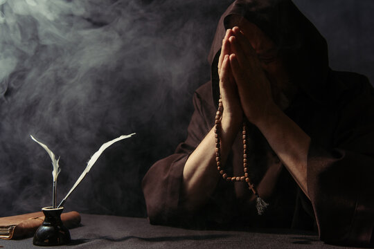 monk with rosary obscuring face with praying hands near inkpot and manuscript on black with smoke.
