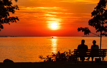 Fototapeta na wymiar Couple sits on park bench in front of setting sun on shore of waterfront park