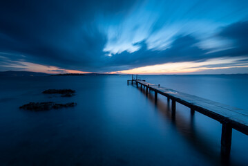 Long exposure photography of pier on the sea, dramatic clouds on skycalm sea with reflection and light on horizon