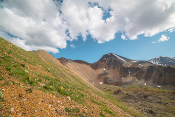 Obraz premium Scenic landscape with sunlit green grassy stone hill and high orange mountain ridge under white clouds in blue sky. Colorful scenery with rocky mountains and sharp rocks in sunlight under cloudy sky.