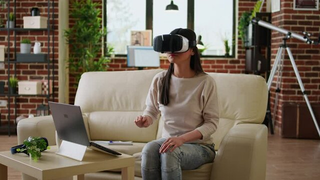 Woman wearing virtual reality headset scrolling through metaverse cyberspace while sitting at home on couch. Young adult person wearing futuristic VR goggles while interacting with cyber reality.