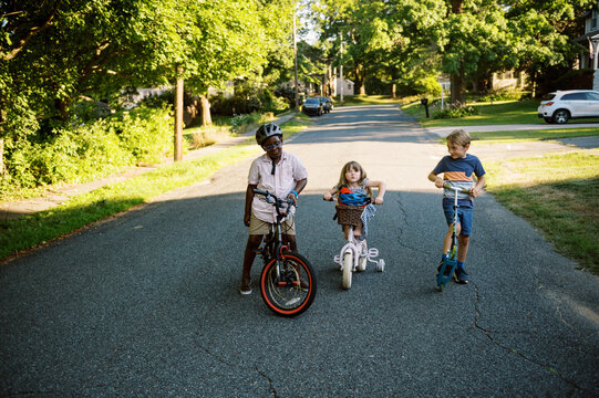 Three Kids On Bikes And Scooter On A Neighborhood Street