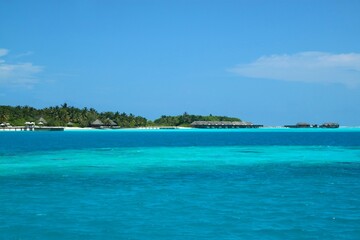 Turquoise water in the Maldives, resort bungalows with thatched-roofs, crystal clear water, blue skies and vegetation   