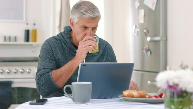 Morning, man working and computer work of a senior male in a home kitchen. Elderly pension person with technology, internet and web email drinking orange juice thinking and reading online information