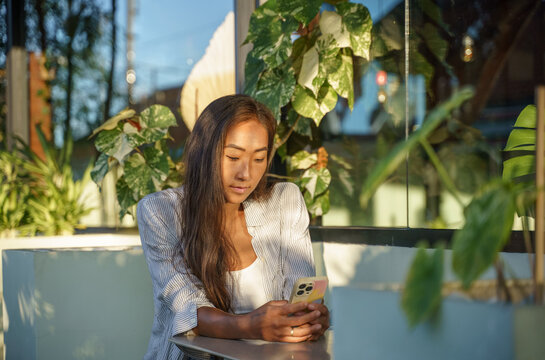 Asian Woman Sitting Alone Using Smartphone In Well Lit Cafe