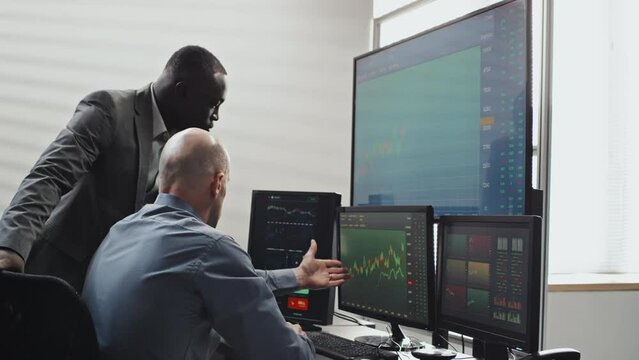 Mature Caucasian man and his young Black colleague looking at stock trading stats on computer monitors and discussing volatility