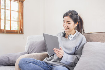 Fototapeta na wymiar Beautiful Asian woman using digital tablet while headphones sitting on sofa at home