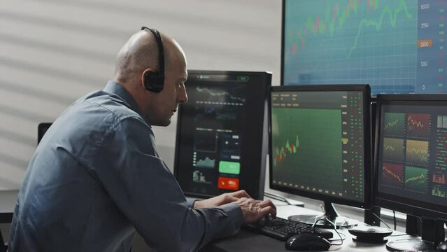 Medium shot of modern mature stock trading specialist wearing headphones with microphone working with data and discussing something with client or colleague