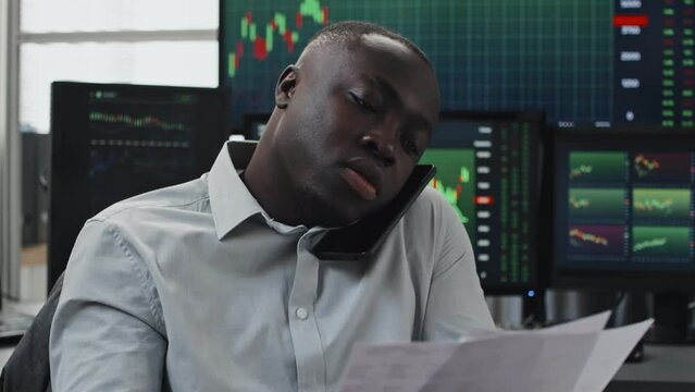 Medium close-up tilt-up of young African American man working as stock trader sitting against computer monitors talking on phone with his client