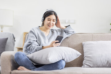 Fototapeta premium Photo of modern asian woman wearing headphones holding cell phone while lying on sofa in bright apartment