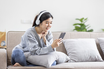 Photo of modern asian woman wearing headphones holding cell phone while lying on sofa in bright...