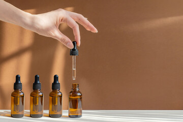 Woman's hand holds pipette with serum over an open bottle on light brown background, in rays of sunlight. Unmarked containers with cosmetics close-up. Open, closed bottles with oil. Concept of mockup - obrazy, fototapety, plakaty