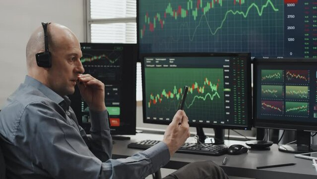 Modern mature Caucasian man wearing headset holding smartphone looking at stock trading charts on screen having conversation with client