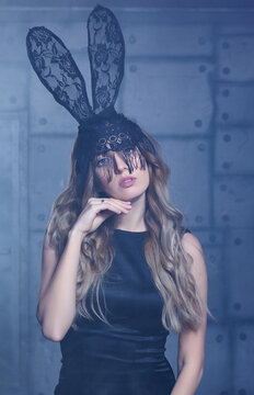 Young blonde woman in black rabbit or hare fancy mask and black dress. Female on smoke and metal wall background.