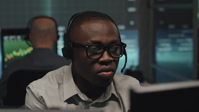 Medium close-up od African American man wearing eyeglasses and headphones with mic working on computer in office late in evening