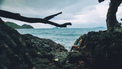 Sexy hot woman meditates, relaxes on rock crack reef hill in stormy morning cloudy sea. Girl in blue swimsuit. Concept feminine, sexual vaginal health, hygiene, womanly, freedom, freshness, recovery