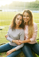 Two women are sitting on the grass in the park next to the football field.