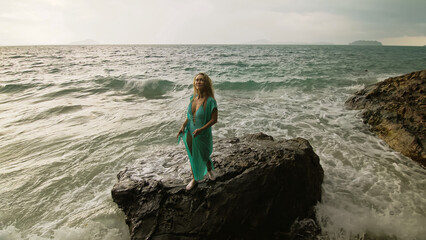 Seductive long-haired woman walks on rock of sea reef stone, stormy cloudy ocean. Woman in blue swimsuit dress tunic. Concept rest in sea, tropical resort coastline traveling tourism summer holidays